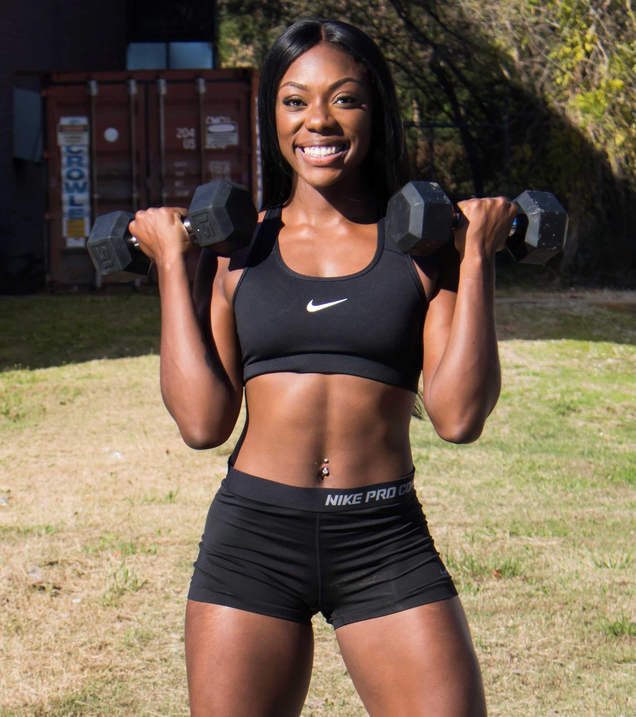 Coach smiling and holding dumbbell's up, dressed in athletic gear.