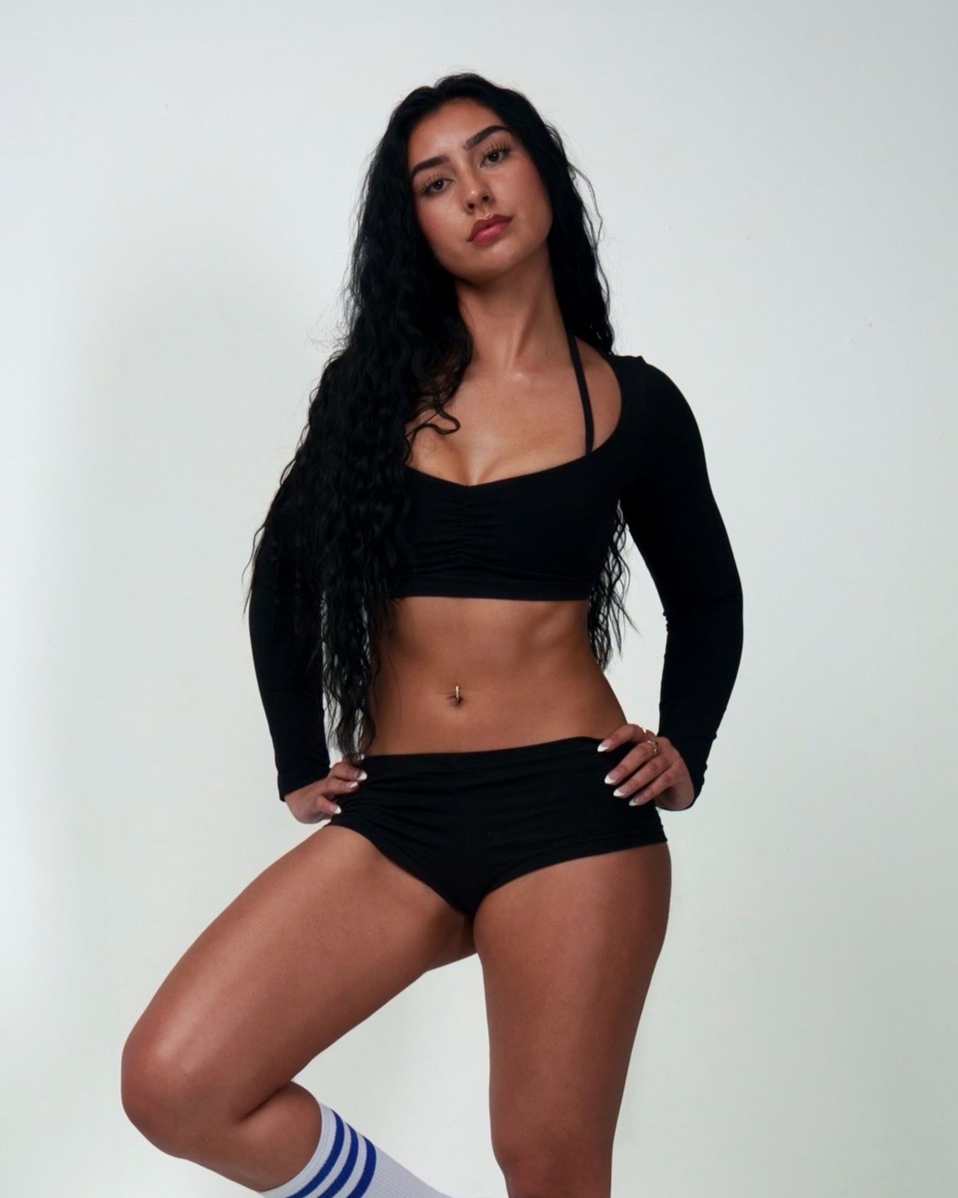 The coach stands against a white background in a black crop top and shorts, hands on her hips.