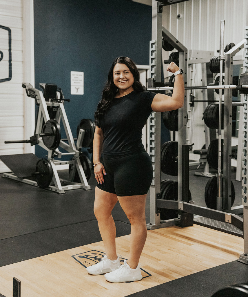 Coach is posing in a black workout outfit with big smile and flexing her arm.